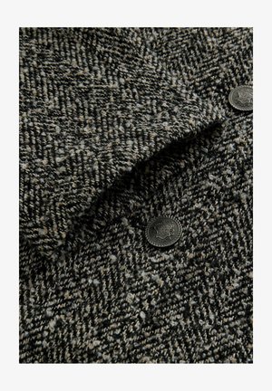 Veste en laine avec un motif en pied de poule texturé noir et beige, ornée de boutons en métal noir décorés de motifs élaborés.