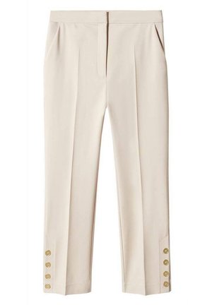 Femme portant un pantalon beige raccourci avec des détails à boutons, une chemise rayée bleue sur un haut gris, des baskets beiges, tenant un sac à main gris.