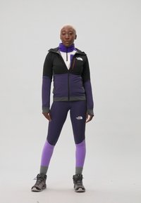 Idrettsantrekket består av en svart og lilla zip-up jakke, lilla leggings og grå tursko med strukturerte såler.