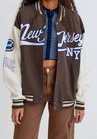 Brązowa i kremowa kurtka varsity z haftowanym napisem 'New Jersey NY', paskowymi ściągaczami przy rękawach i napami, charakteryzująca się miękką teksturą materiału.