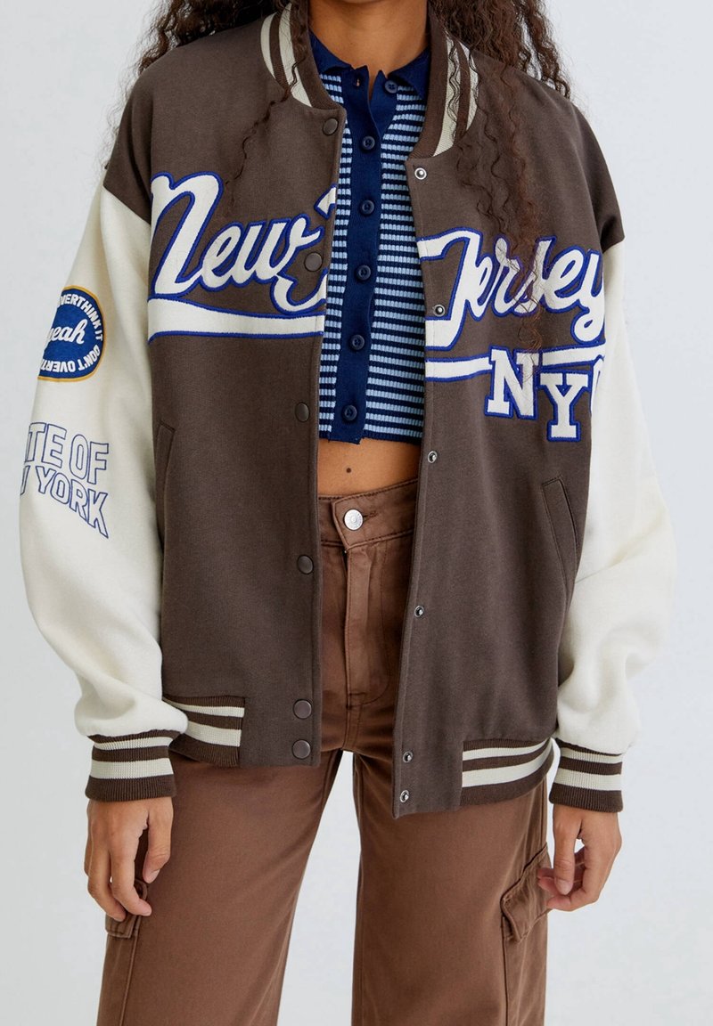 Brązowa i kremowa kurtka varsity z haftowanym napisem 'New Jersey NY', paskowymi ściągaczami przy rękawach i napami, charakteryzująca się miękką teksturą materiału.