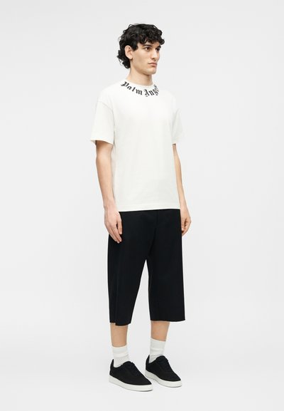 Νέος άνδρας που στέκεται, φορώντας λευκό μπλουζάκι με μαύρο κείμενο στον γιακά, μαύρη φαρδιά βερμούδα μέχρι το γόνατο, λευκές κάλτσες και μαύρα αθλητικά παπούτσια.