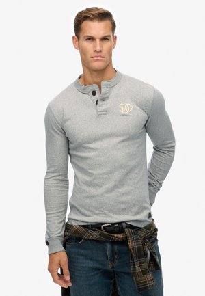 Camisa henley de mangas largas en gris claro con tres botones, que presenta un pequeño logo bordado. Combinada con jeans azules y una camisa a cuadros atada a la cintura.