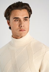 Crèmekleurige coltrui met een gestructureerd patroon, voorzien van een geribde hals en geometrische ontwerpen over het hele kledingstuk. Zacht gebreid materiaal.