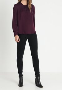 Femme portant un blouse prune à manches longues, un jean skinny noir et des bottines à talons noirs, se tenant contre un fond blanc uni.