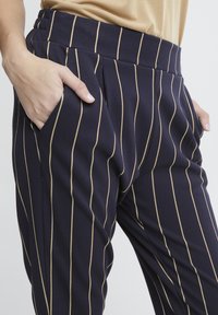 Marineblaue Hosen mit vertikalen cremefarbenen Streifen, ausgestattet mit seitlichen Taschen und einem flachen Bund, gefertigt aus einem glatten, elastischen Stoff.