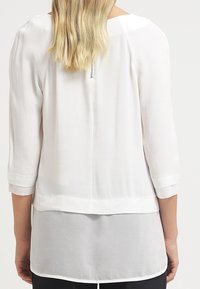 Weißbluse mit Dreiviertelärmeln, geschichtetem Design und einem Reißverschluss auf der Rückseite. Glattes, leichtes Material mit einer leicht durchscheinenden Textur.
