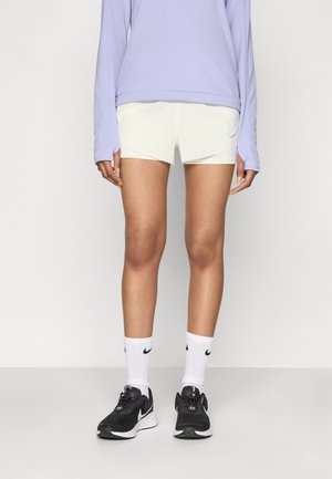 Osoba v světle fialovém dlouhém rukávu, bílých šortkách, bílých ponožkách Nike a černých běžeckých botách Nike, stojící na jednobarevném pozadí.