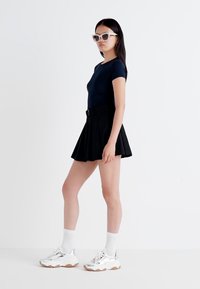 Eine junge Frau steht im Profil, trägt ein enges schwarzes Oberteil, einen ausgestellten schwarzen Rock und weiße Socken mit klobigen Turnschuhen. Sie trägt große Sonnenbrillen.