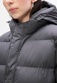Chaqueta acolchada gris con capucha ajustable y diseño acolchado. Textura suave con costuras visibles y un cuello alto. Pendiente plateado visible.