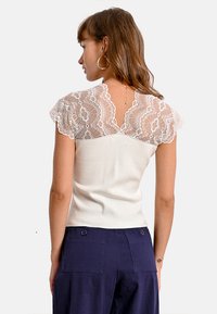 Top bianco aderente con maniche in pizzo trasparente, caratterizzato da un design a V sulla schiena. Il tessuto ha una texture liscia.