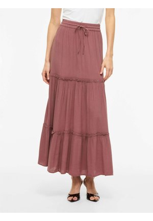 Kvinne iført et støvrosa lang skjørt med lagvis design og snøring i midjen, samt svarte sko med åpen tå, stående mot en hvit bakgrunn.