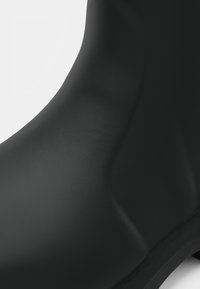 Botín de tobillo de goma negro con una textura suave, que presenta una punta redondeada y un diseño minimalista. Sin herrajes ni patrones visibles.