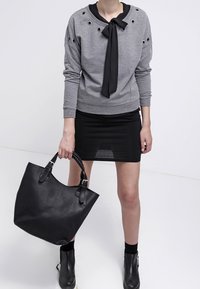 Person som bär en grå tröja med hål, svart slips, svart kjol, ankelstövlar och håller en stor svart läderhandväska.