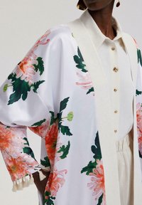 Blommig kimonojacka i vit satin, med stora rosa och gröna blommönster, öppen front med beige kant, samt fransdetaljer på ärmarna.