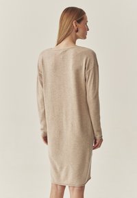 Pletené béžové šaty, dlouh�é rukávy, uvolněný střih, kulatý výstřih, střední délka, hladká textura, minimalistický design. Žádné viditelné vzory ani kovové prvky.