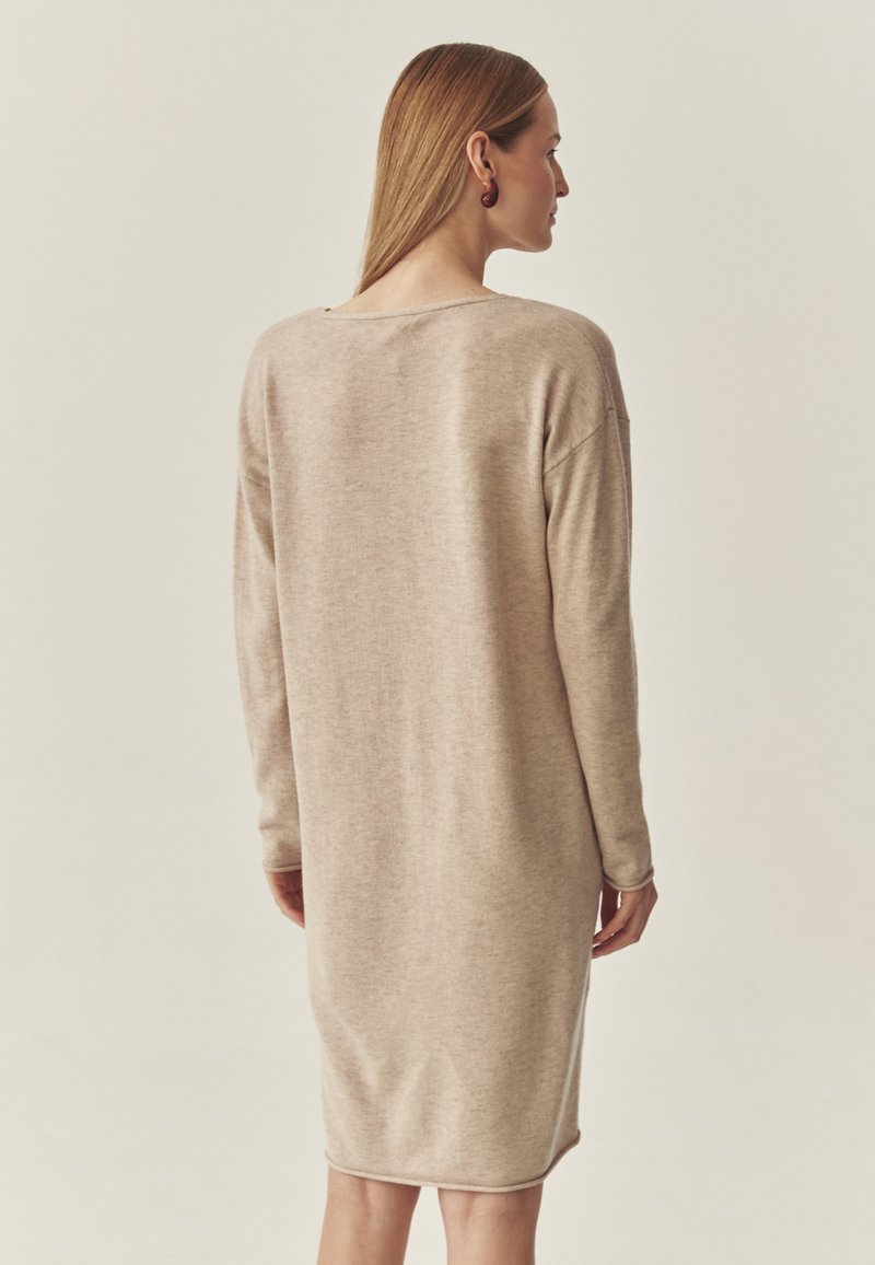 Beżowa sukienka z dzianiny, długie rękawy, luźny krój, okrągły dekolt, midi długość, gładka tekstura, minimalistyczny design. Brak widocznych wzorów lub elementów metalowych.