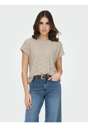 Kvinde med skulderlangt brunt hår iført beige kortærmet bluse med hulmønster, stoppet ned i blå jeans med et mørkebrunt bælte, hånd i lommen.