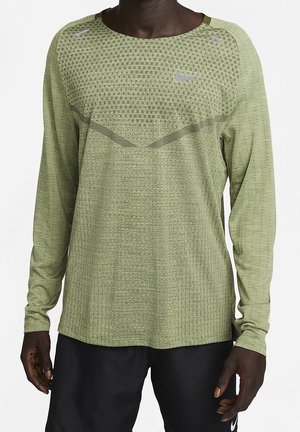 Personne à la peau foncée portant un t-shirt Nike vert clair texturé à manches longues avec un pantalon noir, debout sur fond blanc.