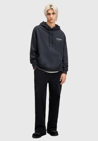 Felpa con cappuccio grigio scuro caratterizzata da una tasca frontale e un logo bianco sul petto, abbinata a pantaloni neri e scarpe da ginnastica scure.