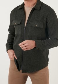 Chaqueta de camisa de mezcla de lana de color verde oscuro con cierre de cremallera, dos bolsillos en el pecho y detalles de botones. Combinándola con una capa base beige y pantalones marrones.