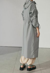 Lichtgrijze, lange jas met capuchon en elastische details aan de mouwen, gedragen over crèmekleurige, taps toelopende broek en zwarte plateauzandalen.