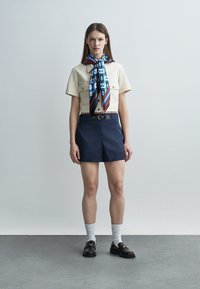 Mujer con camisa beige de manga corta, pantalones cortos de talle alto azul marino con cinturón, bufanda estampada en azul y granate, calcetines blancos y mocasines negros.