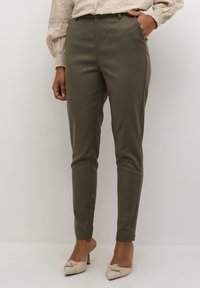 Pantalons fuselés vert olive avec une texture lisse, taille haute et plis cousus, associés à un haut beige et des chaussures à talons beige clair.