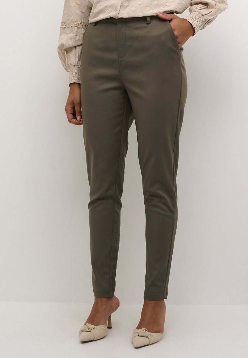 Pantalons fuselés vert olive avec une texture lisse, taille haute et plis cousus, associés à un haut beige et des chaussures à talons beige clair.