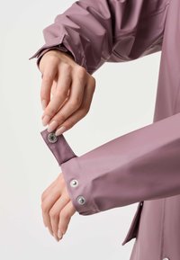 Hænder justerer trykknappen på manchetterne på en støvet lilla regnfrakke mod en ensfarvet hvid baggrund.