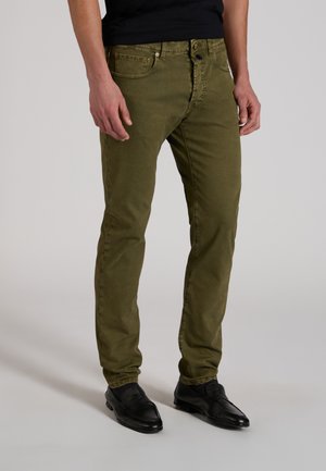 Mężczyzna w dopasowanych oliwkowozielonych spodniach i czarnych eleganckich butach, stojący na jasnym, jednolitym tle z rękami swobodnie opuszczonymi wzdłuż ciała.