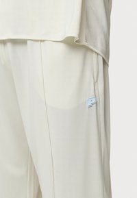 Helle Sporthosen aus glattem Stoff, mit einer Seitentasche und einem blauen „P“-Logo auf dem linken Oberschenkel.