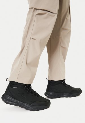 Beżowe spodnie noszone z czarnymi, wysokimi, wodoodpornymi trampkami z grubymi podeszwami i czarno-białymi sznurowadłami, na białym tle.