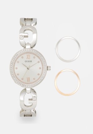 Montre en métal argenté avec un cadran rond, un boîtier en cristal, des accents en or rose et deux cadrans interchangeables : argent et or rose.