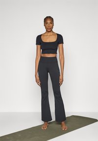 Čierny krátky top so štvorcovým výstrihom, v kombinácii s čiernymi legínami s vysokým pásom a rozšírenými nohavicami. Modelka stojí bosá na zelenej jogovej podložke.