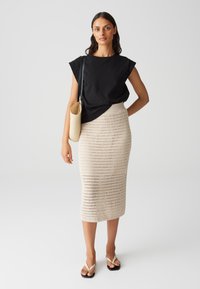 Schwarzes Oberteil mit strukturierten Ärmeln, cremefarbene Häkelröcke und schwarze Flip-Flops. Kleine runde Tasche hängt von der Schulter. Einfaches Design.