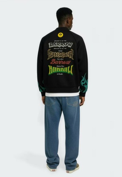 Mężczyzna stojący tyłem, ubrany w czarną kurtkę z kolorowymi napisami "Barrow" na plecach oraz zielonymi wzorami płomieni na rękawach, niebieskie dżinsy i trampki.