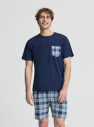 Giovane uomo sorridente, indossa una maglietta blu navy con taschino a quadri e pantaloncini coordinati a quadri su uno sfondo chiaro e uniforme.