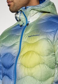 Πουπουλένιο μπουφάν με διαβαθμισμένες αποχρώσεις του μπλε και του πράσινου, με κουκούλα. Διαθέτει πλήρες φερμουά ρ, ρυθμιζόμενα κορδόνια και λογότυπο της μάρκας.