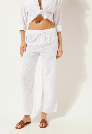 Top de lino blanco atado en la cintura, combinado con pantalones de lino blanco sueltos que presentan una sutil textura floral. Sandalias marrones con tiras completan el look.