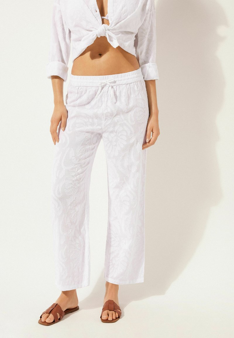 Haut en lin blanc noué à la taille, associé à un pantalon blanc en lin ample avec une subtile texture florale. Des sandales brunes à lanières complètent le look.