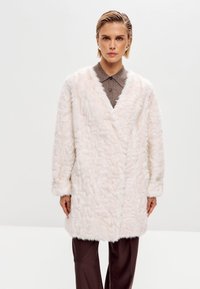 Abrigo de pelo sintético en rosa suave con escote en forma de V, mangas largas y superficie texturizada. Llevado sobre una camisa de botones marrón y pantalones marrones.