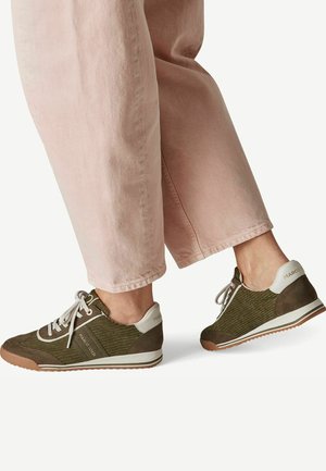 Persoon die olijfgroene en witte sneakers met veters draagt en lichtroze broek tegen een effen achtergrond.