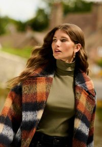 Manteau en laine à carreaux orange, bleu et beige avec des revers surdimensionnés, porté par-dessus un col roulé côtelé olive. Le mannequin a de longs cheveux ondulés.