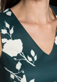 Ärmlös klänning i mörk teal med ett vitt blommönster. Har en V-ringning och en mjuk, slät tygstruktur.