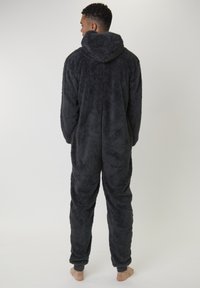 Homme debout pieds nus, tourné le dos, portant une combinaison à capuche grise foncée en peluche, avec des poignets et chevilles à revers.