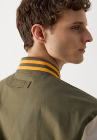 Veste bomber vert olive avec des rayures jaunes et vert foncé sur le col. Présente une étiquette au dos, près du cou. Texture lisse.