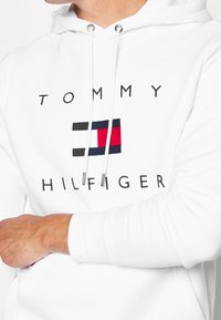 Sudadera blanca con un gran logo bordado de Tommy Hilfiger. Incluye franjas horizontales en rojo, blanco y azul y capucha con cordones.