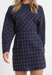 Mörkblårutig klänning med långa ärmar, som har en avskuren midja och utsvängd kjol. Tillverkad av texturerad tyg med vit rutmönster.