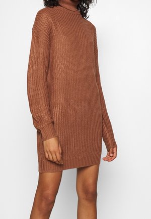 Femme portant une robe pull à col roulé en tricot marron à manches longues, debout devant un fond blanc uni.
